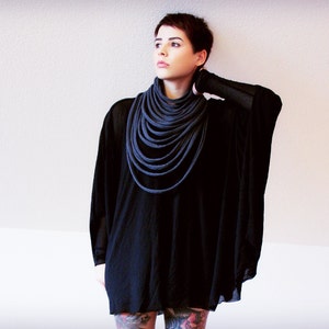 May include: A woman wearing a black tunic with a long, dark grey scarf wrapped around her neck. The scarf is made of multiple layers of fabric.