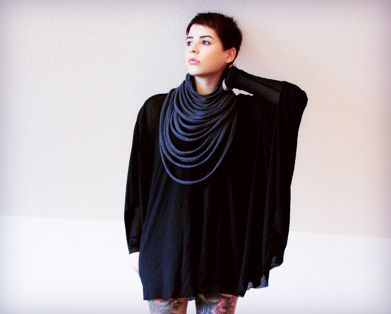 May include: A woman wearing a black tunic with a long, dark grey scarf wrapped around her neck. The scarf is made of multiple layers of fabric.