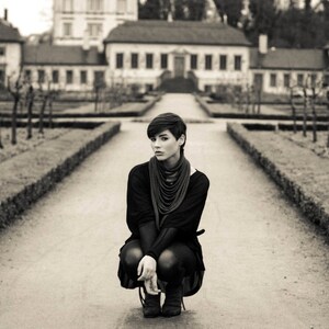 May include: A young person wearing a black sweater and black trousers sits on a path in front of a building. The person is looking at the camera. The building is a large, old house with a white facade and a brown roof.