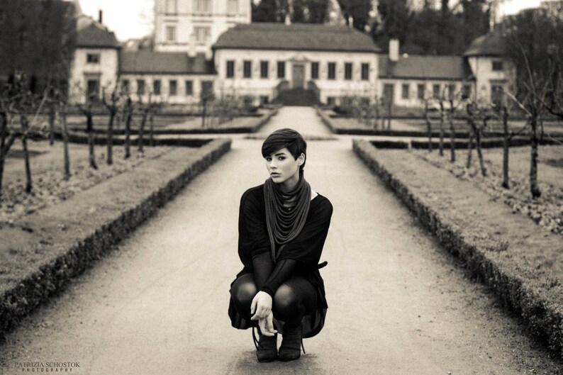 May include: A young person wearing a black sweater and black trousers sits on a path in front of a building. The person is looking at the camera. The building is a large, old house with a white facade and a brown roof.