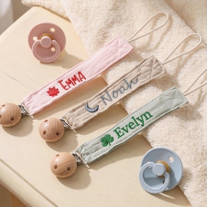 Può includere: Tre clip portaciuccio in tessuto nei colori rosa chiaro, beige e verde salvia, ciascuna ricamata con i nomi Emma, Noah ed Evelyn e piccoli simboli, fissate a clip rotonde in legno, con due ciucci in silicone a tinta unita.
