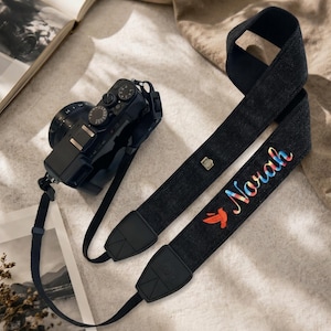 May include: A black camera strap featuring a small metallic camera icon and the name Norah embroidered in a vibrant rainbow gradient, accompanied by a small red bird graphic. The strap is attached to a black digital camera with a lens.