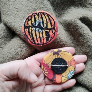 Peut inclure: Deux badges ronds en tissu. L'un présente les mots "Good Vibes" en lettres noires pailletées sur un fond floral rouge et orange. L'autre badge a un motif floral orange, rouge et jaune.