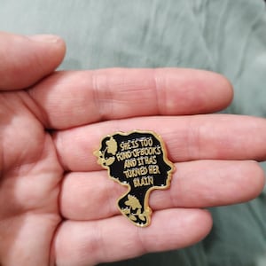 Peut inclure: Une broche en émail noir en forme de profil de tête humaine avec des contours en métal doré. La broche porte le texte 'SHE IS TOO FOND OF BOOKS AND IT HAS TURNED HER BRAIN' en lettres dorées, agrémenté de petits motifs floraux dorés.