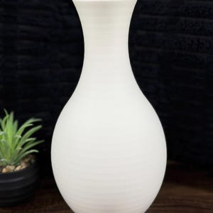 May include: A white ceramic vase with a rounded body and a flared neck. The vase has a textured, ribbed surface and stands on a wooden surface. A small potted plant is visible in the background.
