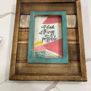 May include: A rectangular wall art piece featuring a dark brown stained wooden frame with horizontal slats. A smaller, distressed turquoise blue frame is centered over the slats, enclosing a white background with pink and yellow fabric accents and the black text "with God all things are possible."