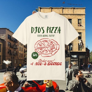 Könnte beinhalten: Cremefarbenes T-Shirt mit einem Pizza-Motiv und dem Text "DJO'S PIZZA" in Grün. Die Grafik zeigt eine Pizza mit roter Soße und Peperoni. Das Shirt hat auch den Text "PIZZA BEING PIZZA!" und "Order now!"