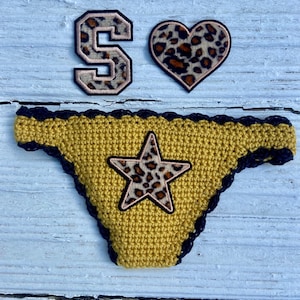 May include: A mustard yellow crochet bikini bottom with a dark navy blue scalloped trim and a central star-shaped patch featuring a brown and black leopard print. Above it are two matching leopard print patches, one shaped like the letter S and one heart.