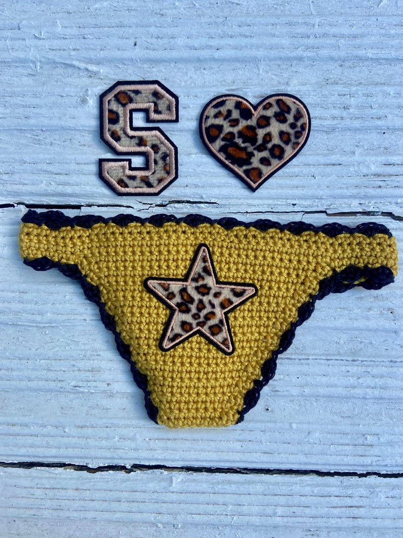 May include: A mustard yellow crochet bikini bottom with a dark navy blue scalloped trim and a central star-shaped patch featuring a brown and black leopard print. Above it are two matching leopard print patches, one shaped like the letter S and one heart.
