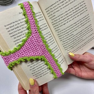 May include: A handmade crochet bookmark featuring a pink center with a bright green scalloped edge. The bookmark is shaped like a pair of underwear and is draped over the open pages of a book titled The Gunpowder and Glory Girls.