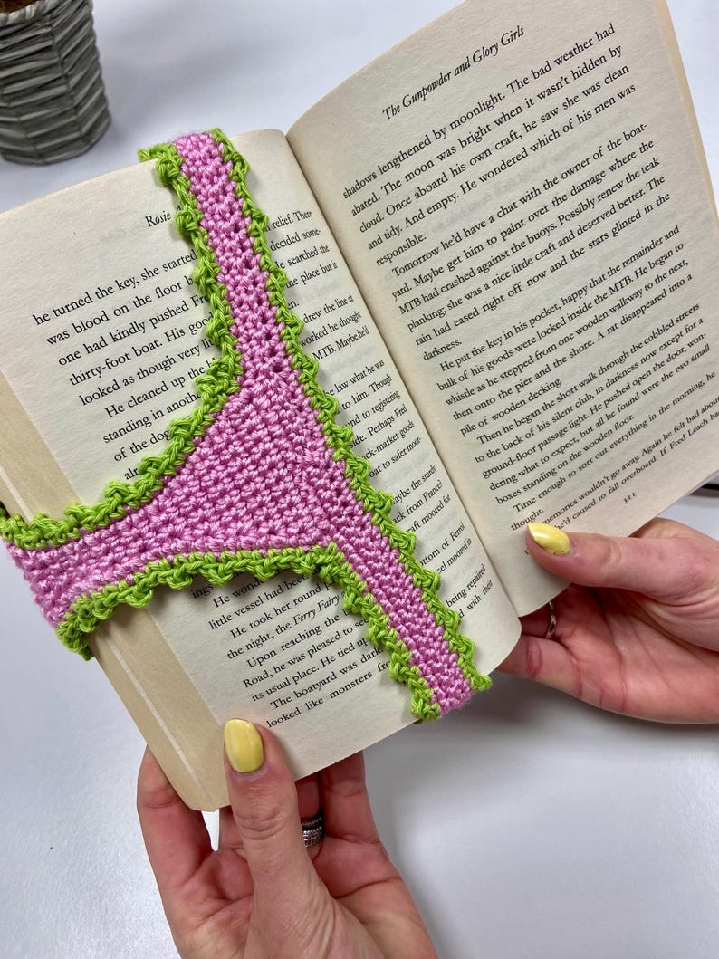 May include: A handmade crochet bookmark featuring a pink center with a bright green scalloped edge. The bookmark is shaped like a pair of underwear and is draped over the open pages of a book titled The Gunpowder and Glory Girls.