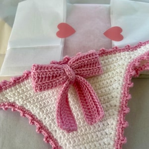 May include: A white crochet bikini bottom featuring a pink crochet bow centered on the front. The edges of the garment are finished with a pink scalloped crochet trim. The piece is constructed from white and pink yarn in a textured stitch pattern.