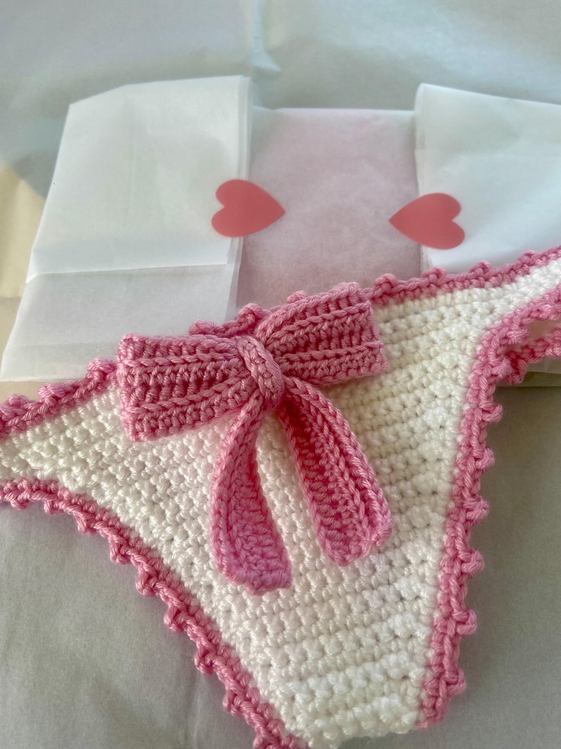 May include: A white crochet bikini bottom featuring a pink crochet bow centered on the front. The edges of the garment are finished with a pink scalloped crochet trim. The piece is constructed from white and pink yarn in a textured stitch pattern.