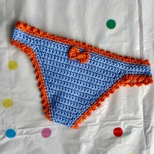May include: A light blue crochet bikini bottom featuring a vibrant orange scalloped trim along the edges and waistband. A small, matching orange crochet bow is centered on the front of the waistband, adding a decorative detail to the piece.