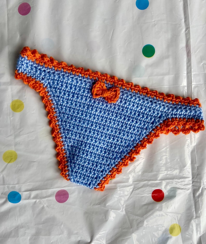 May include: A light blue crochet bikini bottom featuring a vibrant orange scalloped trim along the edges and waistband. A small, matching orange crochet bow is centered on the front of the waistband, adding a decorative detail to the piece.
