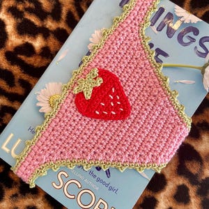 May include: A triangular crochet bandana in light pink with a decorative light green scalloped border. A red strawberry appliqu&eacute; with a light green stem and white embroidered seeds is centered on the fabric. The item is placed on a book.
