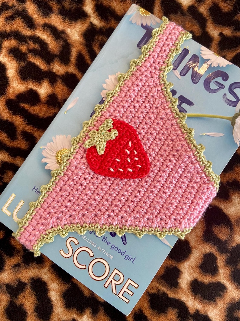 May include: A triangular crochet bandana in light pink with a decorative light green scalloped border. A red strawberry appliqu&eacute; with a light green stem and white embroidered seeds is centered on the fabric. The item is placed on a book.