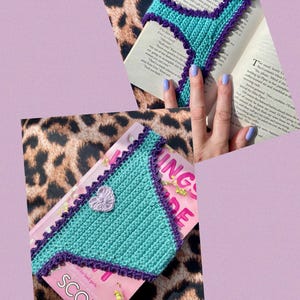 May include: A triangular crochet bookmark in bright teal with a dark purple scalloped border and a small, light purple crocheted heart accent in the center. The bookmark is shown placed on top of an open book with printed text.