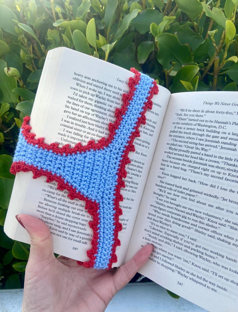 May include: A light blue crocheted bookmark with a scalloped red border, shaped like a pair of underwear, placed inside an open book with visible printed text on the pages. The bookmark features a central light blue panel with red trim.