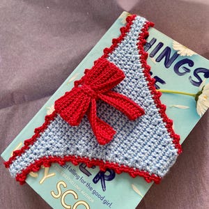 May include: A triangular crochet bookmark in light blue yarn with a decorative red scalloped border and a prominent red crocheted bow attached to the center. The bookmark is placed on top of a book with a light blue cover featuring white daisy graphics.
