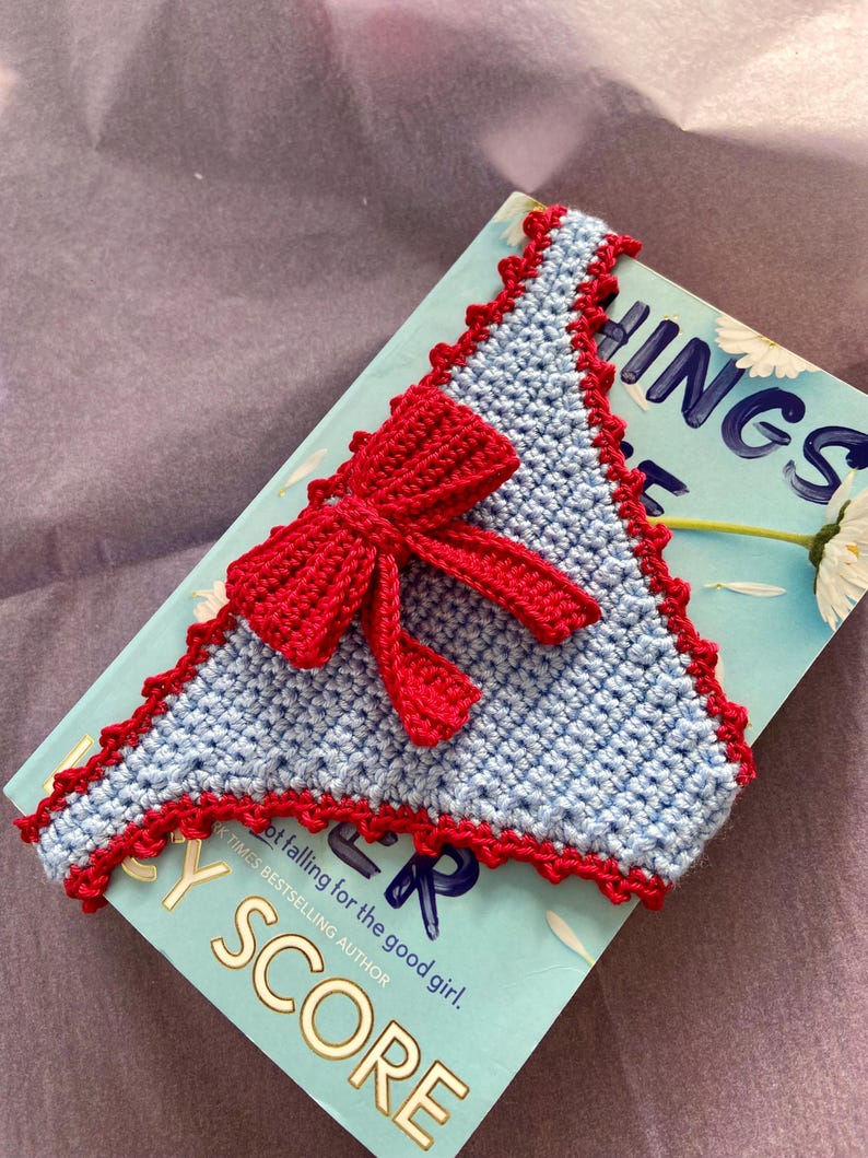 May include: A triangular crochet bookmark in light blue yarn with a decorative red scalloped border and a prominent red crocheted bow attached to the center. The bookmark is placed on top of a book with a light blue cover featuring white daisy graphics.