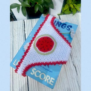 May include: A white triangular crochet bookmark with a red scalloped edge and a central circular watermelon motif featuring a red center, black seeds, and a light green rind, resting on a light blue book cover with the visible text SCORE.