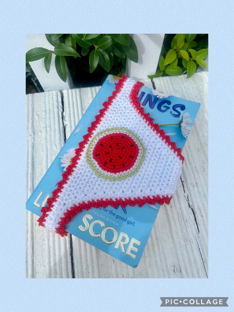 May include: A white triangular crochet bookmark with a red scalloped edge and a central circular watermelon motif featuring a red center, black seeds, and a light green rind, resting on a light blue book cover with the visible text SCORE.