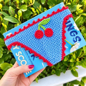 May include: A light blue crochet book sleeve featuring a red scalloped border and a central cherry applique with two red circular fruits and a green leaf. The sleeve is positioned over a blue book cover with white daisy graphics and text.
