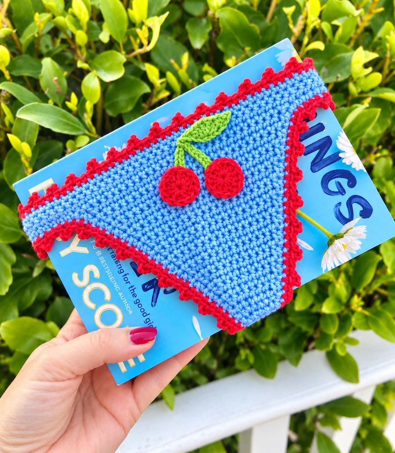 May include: A light blue crochet book sleeve featuring a red scalloped border and a central cherry applique with two red circular fruits and a green leaf. The sleeve is positioned over a blue book cover with white daisy graphics and text.