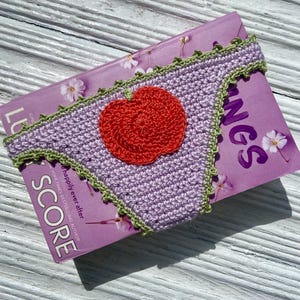 May include: A lavender crochet bikini bottom featuring a bright red apple appliqu&eacute; centered on the front. The edges of the garment are finished with a decorative light green scalloped trim, providing a contrasting border to the purple fabric.