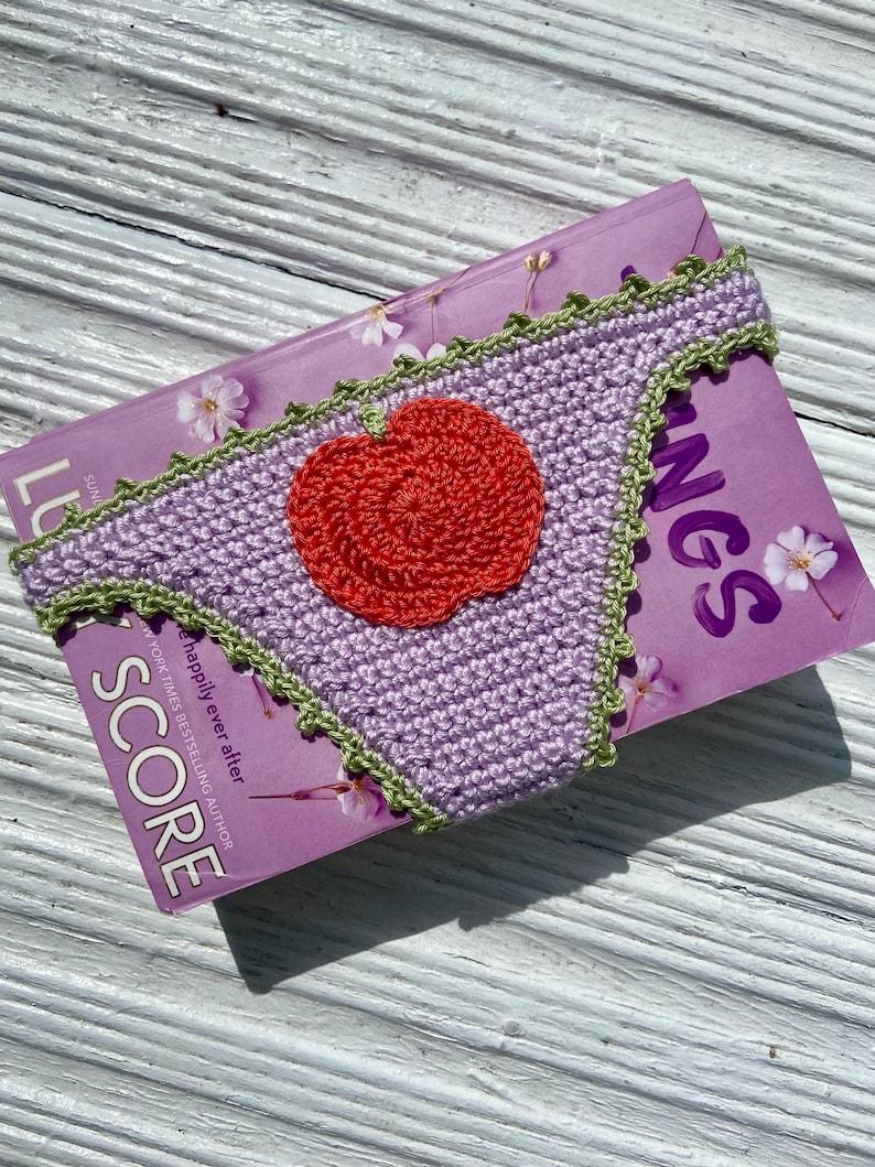 May include: A lavender crochet bikini bottom featuring a bright red apple appliqu&eacute; centered on the front. The edges of the garment are finished with a decorative light green scalloped trim, providing a contrasting border to the purple fabric.