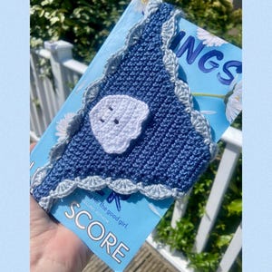 May include: A triangular crochet book sleeve in dark blue with a light blue scalloped border. A white crocheted ghost applique with two black eyes is attached to the center. The sleeve is placed over a light blue book cover with white flowers.