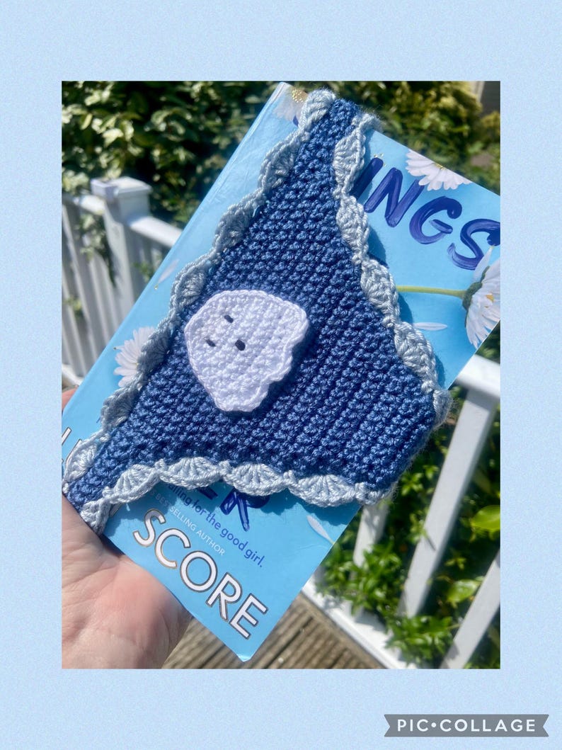 May include: A triangular crochet book sleeve in dark blue with a light blue scalloped border. A white crocheted ghost applique with two black eyes is attached to the center. The sleeve is placed over a light blue book cover with white flowers.