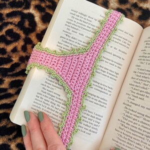 May include: A handmade crochet bookmark featuring a pink central strip with a textured light green scalloped border. The bookmark is shaped like a pair of underwear and is positioned vertically between the pages of an open book with printed text.