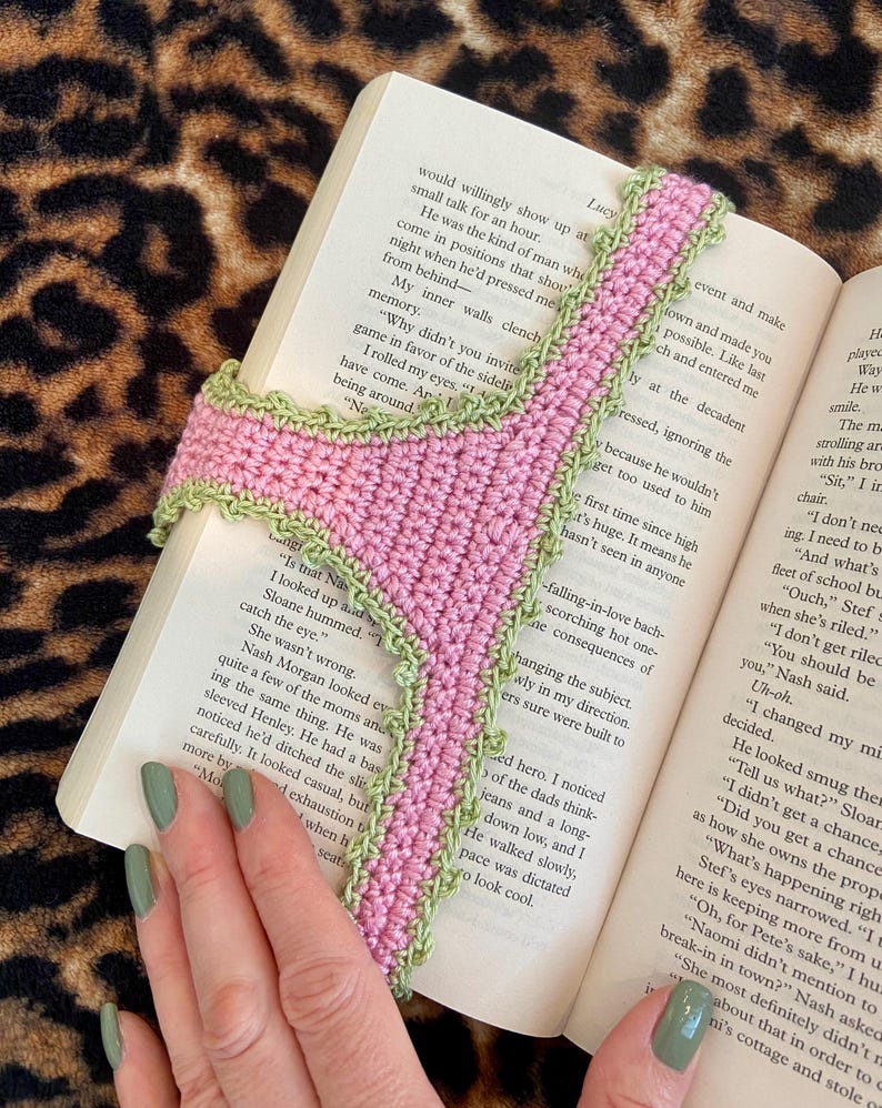 May include: A handmade crochet bookmark featuring a pink central strip with a textured light green scalloped border. The bookmark is shaped like a pair of underwear and is positioned vertically between the pages of an open book with printed text.