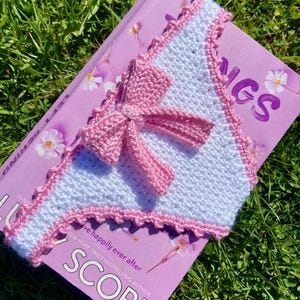 May include: A triangular crochet bookmark featuring a white textured stitch pattern with a pink scalloped border and a central pink crochet bow. The bookmark is placed on a purple book cover with the visible text SCORE and NEW YORK TIMES BESTSELLING AUTHOR.