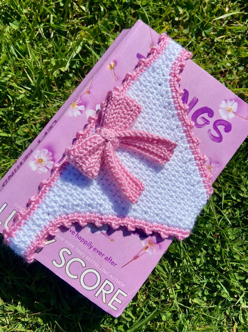 May include: A triangular crochet bookmark featuring a white textured stitch pattern with a pink scalloped border and a central pink crochet bow. The bookmark is placed on a purple book cover with the visible text SCORE and NEW YORK TIMES BESTSELLING AUTHOR.