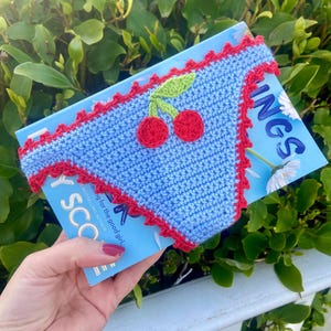 Puede incluir: Una funda de libro de ganchillo azul con ribete rojo y un diseño de cereza. La funda está envuelta alrededor de un libro con el título "Things" visible. El diseño de cereza presenta dos cerezas rojas y hojas verdes.