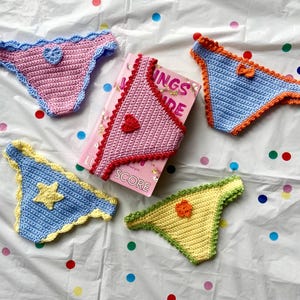 May include: Four handmade crochet bikini bottoms in pink, blue, and yellow with decorative heart, star, and flower appliques, arranged around a pink book titled "INGS IDE SCORE". The crochet pieces feature scalloped edges in contrasting colors.