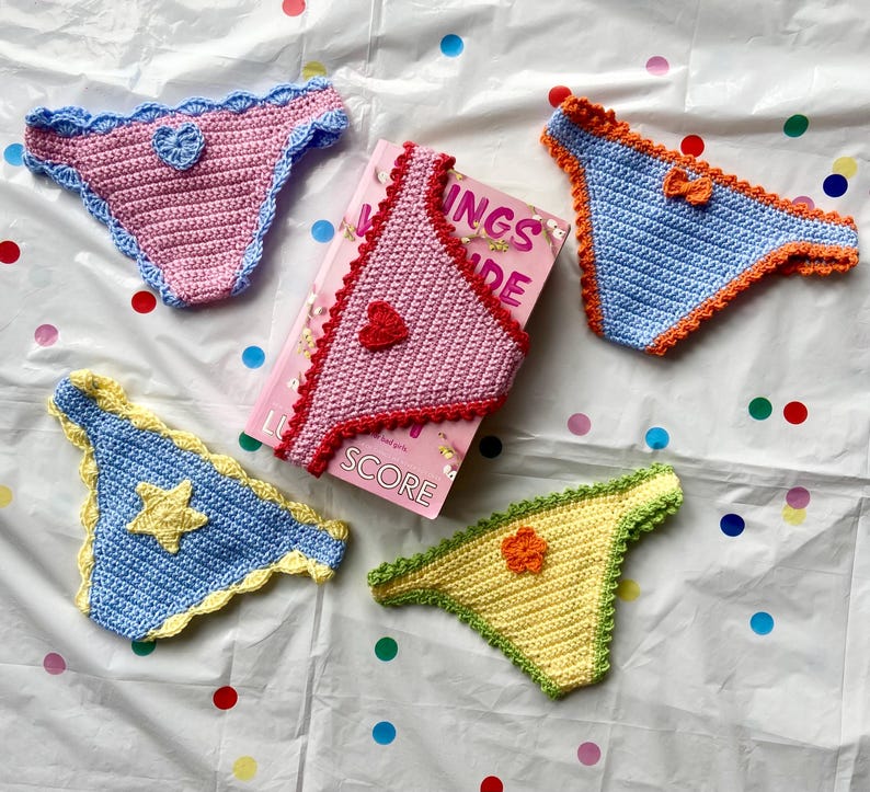 May include: Four handmade crochet bikini bottoms in pink, blue, and yellow with decorative heart, star, and flower appliques, arranged around a pink book titled "INGS IDE SCORE". The crochet pieces feature scalloped edges in contrasting colors.