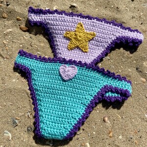 May include: Two crochet bikini bottoms featuring a dark purple scalloped trim. The top piece is light lavender with a centered gold star appliqu&eacute;. The bottom piece is turquoise with a small, light lavender heart-shaped appliqu&eacute; centered at the top.