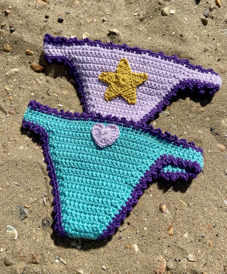 May include: Two crochet bikini bottoms featuring a dark purple scalloped trim. The top piece is light lavender with a centered gold star appliqu&eacute;. The bottom piece is turquoise with a small, light lavender heart-shaped appliqu&eacute; centered at the top.