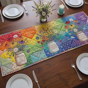 Puede incluir: Un colorido camino de mesa acolchado con un diseño de gradiente de arcoíris. Presenta tarros de cristal bordados llenos de flores vibrantes. Las palabras "LOVE IS LOVE" y "PRIDE" se muestran en el camino de mesa, colocado sobre una mesa de madera.