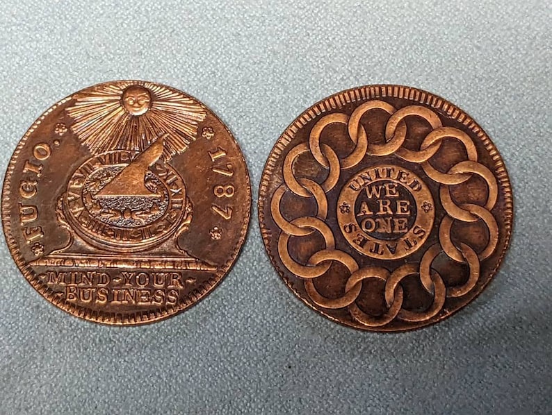 May include: Two copper-colored coins with different designs. The first coin features a sun with rays, a compass, and the text "FUGIO 1787 MIND YOUR BUSINESS". The second coin features a circle of linked rings with the text "UNITED WE ARE ONE STATES".