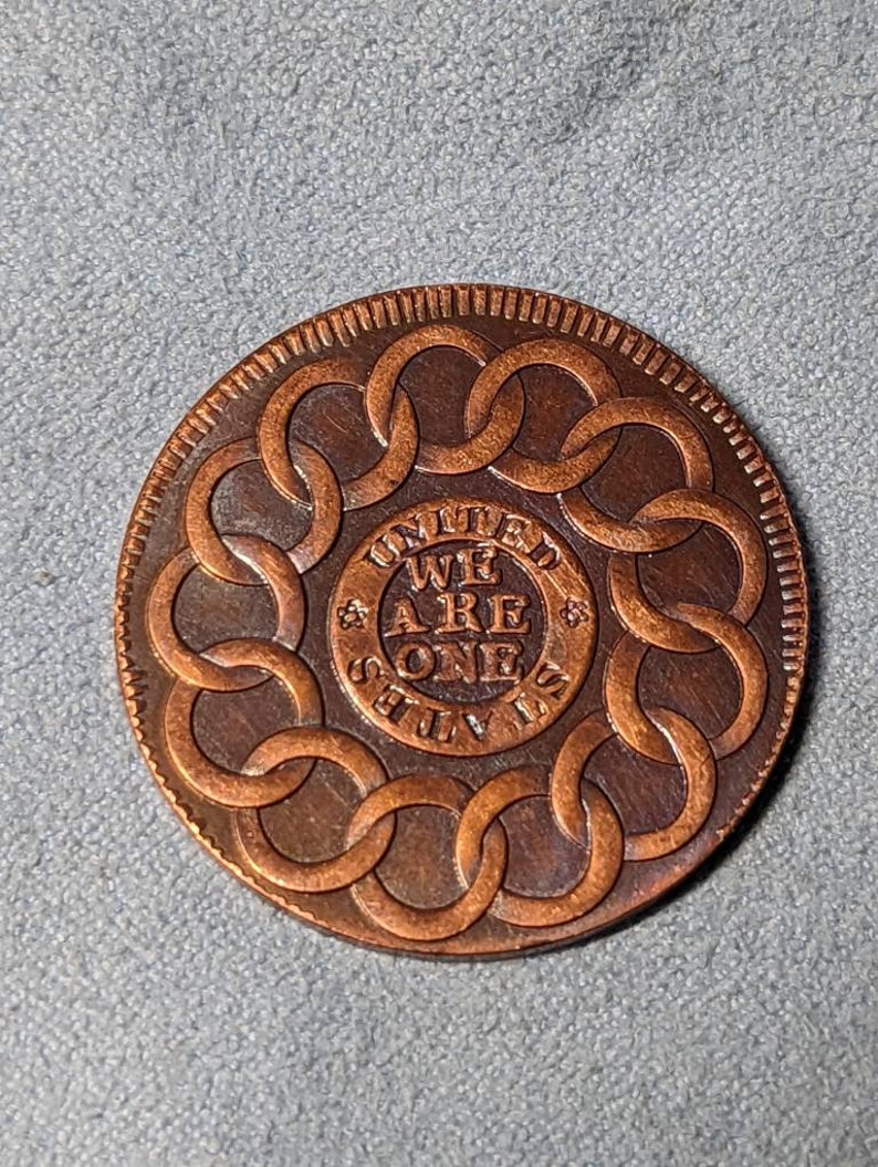 May include: A copper-colored coin with a circular design featuring a chain link pattern around the edge. The center of the coin has the words "UNITED WE ARE ONE STILL" inscribed in a circle.
