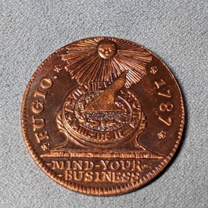May include: A copper-colored coin with a sun and a bird design. The coin reads "FUGIO 1787" and "MIND-YOUR-BUSINESS".