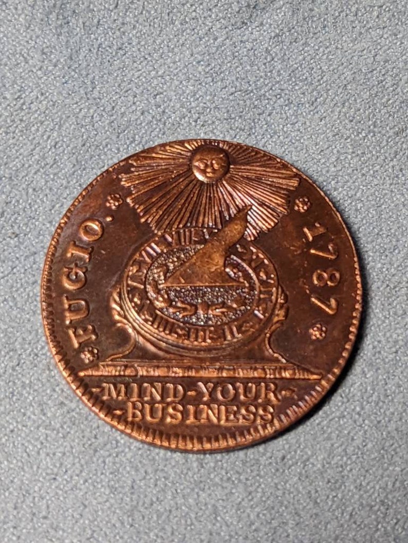 May include: A copper-colored coin with a sun and a bird design. The coin reads "FUGIO 1787" and "MIND-YOUR-BUSINESS".