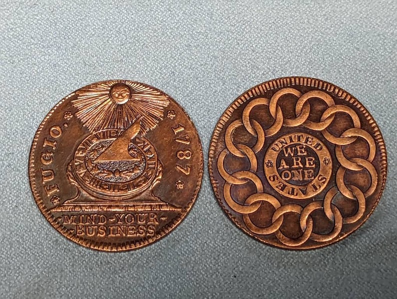May include: Two copper coins with different designs. The first coin features a sun with rays, a compass, and the text "FUGIO 1787 MIND YOUR BUSINESS". The second coin features a circle of linked chains with the text "UNITED STATES WE ARE ONE".