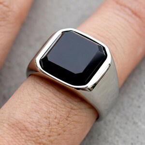 May include: A silver-toned signet ring featuring a smooth, polished, rectangular black stone set into the center of the metal band. The ring has a wide, solid shank that tapers slightly toward the bottom, showcasing a clean, reflective finish.