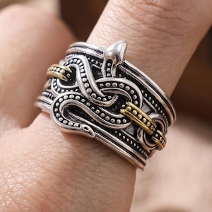 May include: A wide silver-toned ring featuring an intricate, raised snake motif that wraps around the band. The design includes oxidised recessed areas for contrast and two small, gold-toned metallic accents integrated into the snake's body pattern.